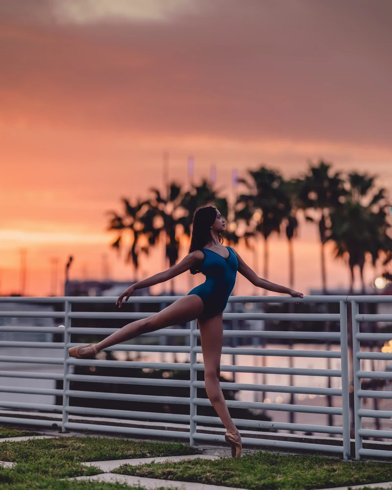 Grace in a blue leotard en pointe beside a waterfront railing at sunset
